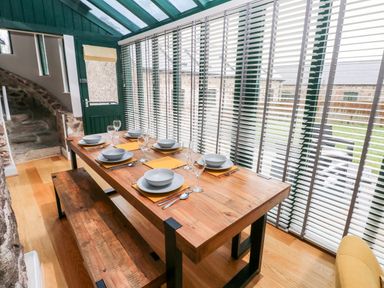 A dining area with a wooden table set for six and large windows with blinds at Skomer Cottage in Sandy Haven near Milford Haven
