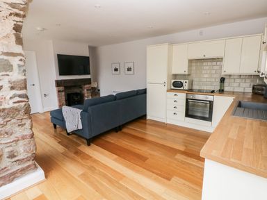 A kitchen and living room area with wooden floors a dark sofa a TV above a brick fireplace and white kitchen cabinets at Skomer Cottage in Sandy Haven near Milford Haven