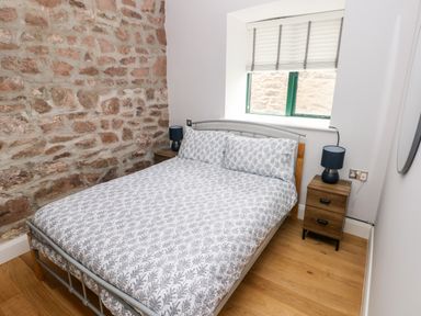 A bedroom with a patterned bed, two bedside tables with lamps, a stone wall, and a window at Skomer Cottage in Sandy Haven near Milford Haven