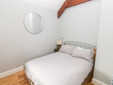A bedroom with a double bed, bedside table with lamp, a round wall mirror, wooden flooring, and wooden ceiling beam at Skomer Cottage in Sandy Haven near Milford Haven