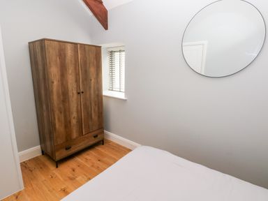 A bedroom corner with a wooden wardrobe a small window with blinds a round mirror and part of a bed at Skomer Cottage in Sandy Haven near Milford Haven
