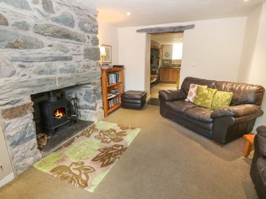 A living room with a stone fireplace and wood stove a leather sofa with cushions a wooden bookshelf and an open doorway leading to a kitchen at Tyn Llwyn in Eisingrug near Harlech