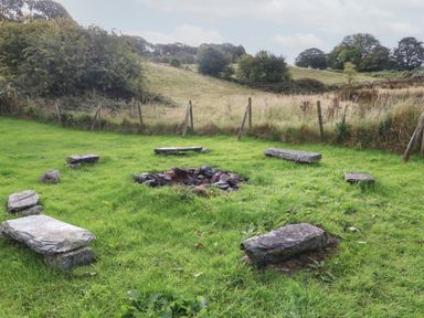 A grassy area with a stone fire pit surrounded by stone benches and a wooden fence in the background at Tyn Llwyn in Eisingrug near Harlech