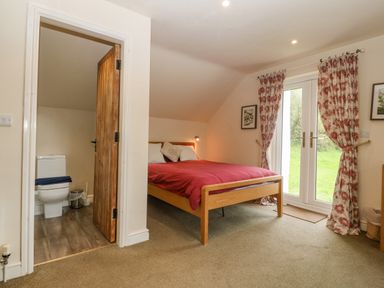 A bedroom with a wooden bed with red bedding next to glass doors with floral curtains and a partially open wooden door revealing a bathroom at Tyn Llwyn in Eisingrug near Harlech