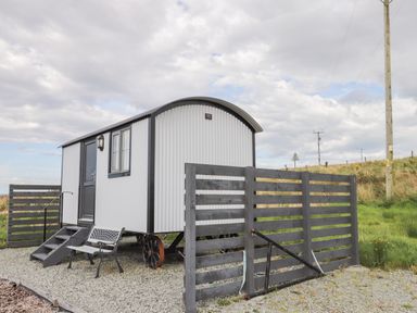 A small white shepherds hut with a black door and window next to a gray wooden fence and bench at Shepherds Hut in Dunvegan