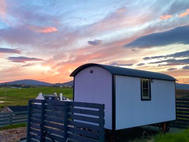 A small white corrugated metal shepherds hut with a window next to a wooden fence with green hills and houses in the background at sunset at Shepherds Hut in Dunvegan