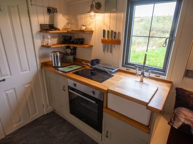 A compact kitchen with wooden countertops a white sink electric hob an oven kettle and shelves with kitchenware at Shepherds Hut in Dunvegan