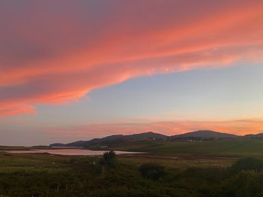 A landscape with hills green fields and a body of water under an orange and pink sky at sunset at Shepherds Hut in Dunvegan