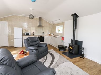 A living room with gray recliner chairs a kitchen area with white cabinets and appliances and a black wood stove at Cliff Barn in Burton Bradstock