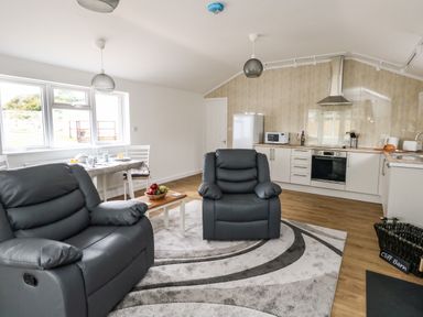 A living room with two gray recliner chairs a small coffee table with fruit a dining table set near windows and kitchen appliances at Cliff Barn in Burton Bradstock