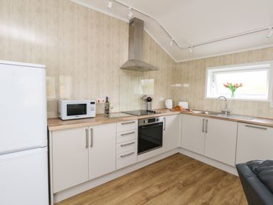 A kitchen with white cabinets a microwave a wine bottle an oven and a sink with flowers on the windowsill at Cliff Barn in Burton Bradstock