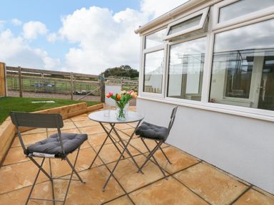 An outdoor patio with a small round table and two chairs with cushions and flowers in a vase at Cliff Barn in Burton Bradstock