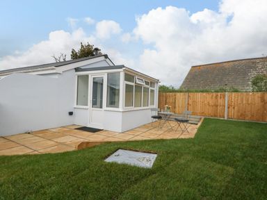 A small patio area with table and chairs next to a white conservatory building and grass lawn at Cliff Barn in Burton Bradstock