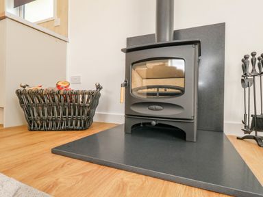 A wood stove with a metal surround on a wooden floor next to a basket with firewood and fireplace tools at Cliff Barn in Burton Bradstock