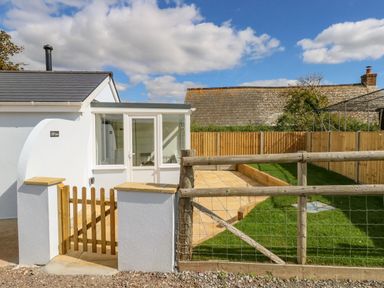 A small enclosed garden with grass and a tiled patio next to a white building with a wooden gate at Cliff Barn in Burton Bradstock