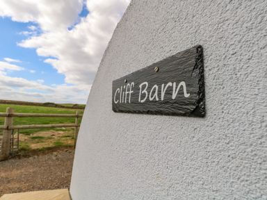 A sign reading Cliff Barn mounted on a white textured wall with a wooden fence and green field in the background at Cliff Barn in Burton Bradstock