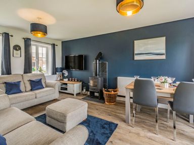 A living room with a beige sofa and ottoman a wood stove a dining table with chairs and a wall-mounted TV at Lantern House in Filey
