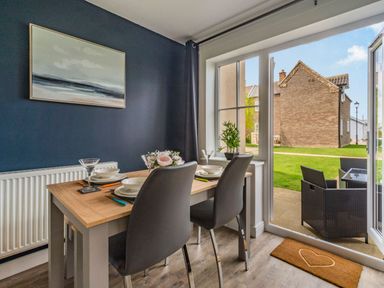 A dining area with a table, four chairs, tableware, a plant, and a painting next to glass doors opening to an outdoor patio at Lantern House in Filey