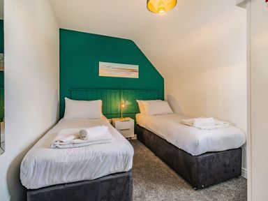 A bedroom with two single beds white bedding and towels a bedside table with a lamp and a green accent wall at Lantern House in Filey