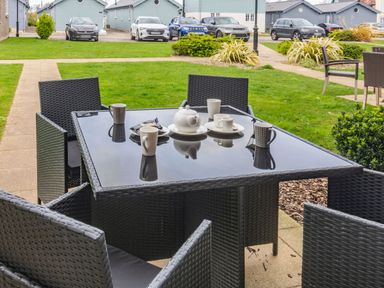 An outdoor patio with a black wicker table and chairs set with cups and a teapot in a grassy area with parked cars and blue houses in the background