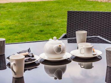 A glass table with a white teapot cups plates and biscuits outside with grass in the background
