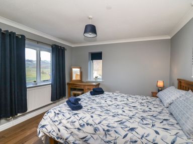 A bedroom with a bed covered with floral bedding a wooden vanity desk and two windows with blue curtains at Pen Y Banc in Llanfaredd near Builth Wells