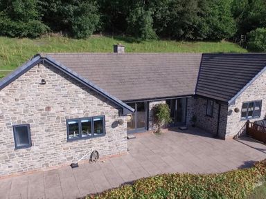 Stone house with tiled roof and windows overlooking a paved patio with grass and trees behind at Pen Y Banc in Llanfaredd near Builth Wells