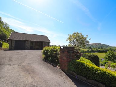 A driveway leading to a stone building with a garage and bench surrounded by greenery at Pen Y Banc in Llanfaredd near Builth Wells
