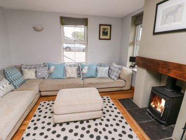 A living room with a corner sofa and cushions a polka dot rug a wood burning stove and windows at Cae Du in Dwyran near Niwbwrch Newborough