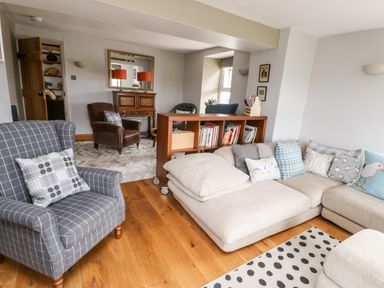 A living room with a beige sectional sofa with cushions a gray checkered armchair a wooden bookshelf on wheels and a brown armchair at Cae Du in Dwyran near Niwbwrch Newborough