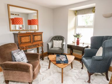 A sitting room with three armchairs a wooden coffee table an antique wooden cabinet with two orange lamps and a mirror above it at Cae Du in Dwyran near Niwbwrch Newborough
