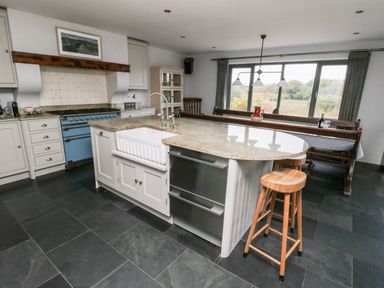 A kitchen with an island counter, wooden stools, blue stove, and a dining area with a bench and large window at Cae Du in Dwyran near Niwbwrch Newborough