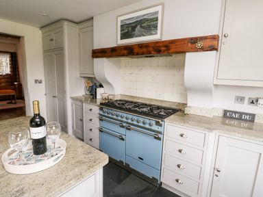 A kitchen with a blue stove white cabinets a bottle of wine with wine glasses on a tray and a picture above the stove at Cae Du in Dwyran near Niwbwrch Newborough