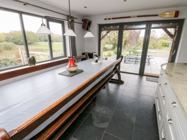 A dining area with a long wooden table and bench seating near large windows and glass doors at Cae Du in Dwyran near Niwbwrch Newborough