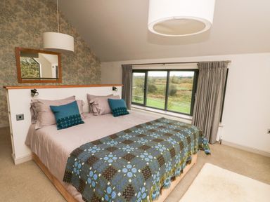 A bedroom with a bed covered with a patterned blanket and pillows next to a window with curtains at Cae Du in Dwyran near Niwbwrch Newborough