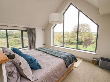 A bedroom with a bed facing a large triangular window overlooking trees and a circular light fixture hanging from the ceiling at Cae Du in Dwyran near Niwbwrch Newborough