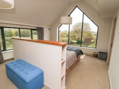 A bedroom with a bed by a large triangular window and a blue bench near a half wall at Cae Du in Dwyran near Niwbwrch Newborough