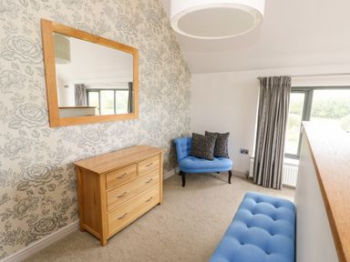 A room with a wooden dresser and mirror blue seating and a large window with grey curtains at Cae Du in Dwyran near Niwbwrch Newborough