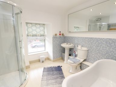 A bathroom with a shower enclosure, sink, toilet, window with floral blind, and a bathtub at Cae Du in Dwyran near Niwbwrch Newborough