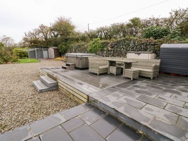 An outdoor patio area with wicker furniture table chairs hot tub storage sheds and gravel ground at Cae Du in Dwyran near Niwbwrch Newborough
