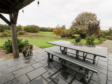 An outdoor patio with a picnic table and two wooden bear sculptures near green lawn and trees at Cae Du Dwyran near Niwbwrch Newborough