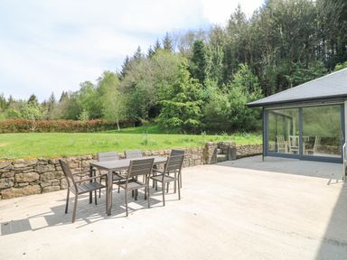 An outdoor patio with a table and six chairs next to a stone wall and a building with glass sliding doors surrounded by trees at Coffey's Ballymacarbry County Waterford