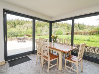 A dining area with a wooden table and six chairs surrounded by large glass windows showing an outdoor patio and green garden at Coffey's in Ballymacarbry County Waterford