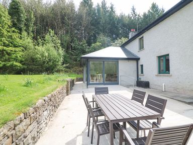 An outdoor patio with a wooden table and chairs beside a house with large glass doors and surrounded by trees