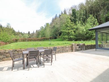 An outdoor patio with a dining table and six chairs near a stone wall and green lawn surrounded by trees at Coffey's in Ballymacarbry County Waterford
