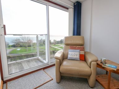 A beige armchair with two pillows near a wooden side table with a cup and book next to a glass sliding door overlooking a garden and sea at Kiama in Weymouth