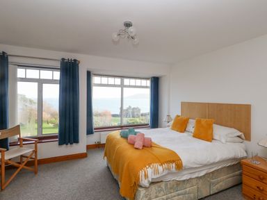 A bedroom with a bed, wooden headboard, bedside table with lamp, chair, blue curtains, and large windows showing ocean view at Kiama in Weymouth