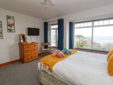 A bedroom with a bed, chest of drawers, chair, wall-mounted TV, blue curtains and large windows overlooking a garden and the sea at Kiama in Weymouth