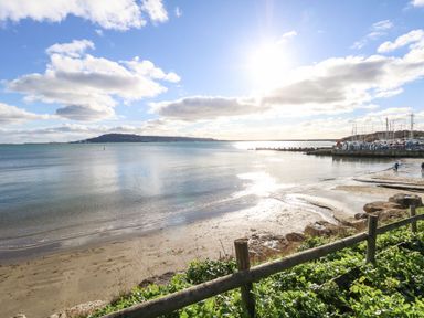 A beach with a wooden fence and green plants near a marina with boats under a clear sky at Kiama in Weymouth