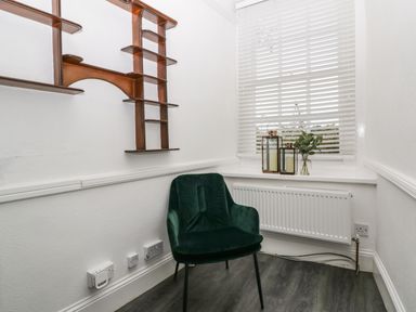 A small room with a green chair wooden wall shelves and a window with white blinds and candles at Alcombe House Minehead